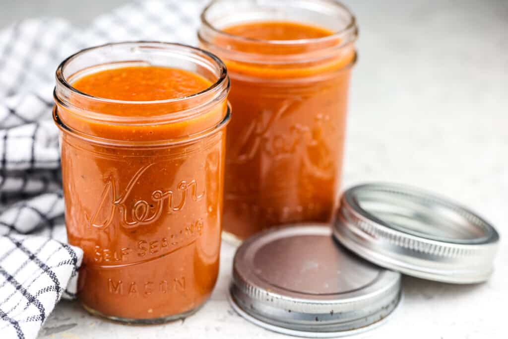 Two mason jars filled with red sauce next two silver lids on a white backdrop with a checkboard white and black towel.