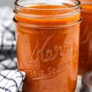 Picture of mason jar holding red enchilada sauce with second jar in the back next to two silver lids and a black towel.