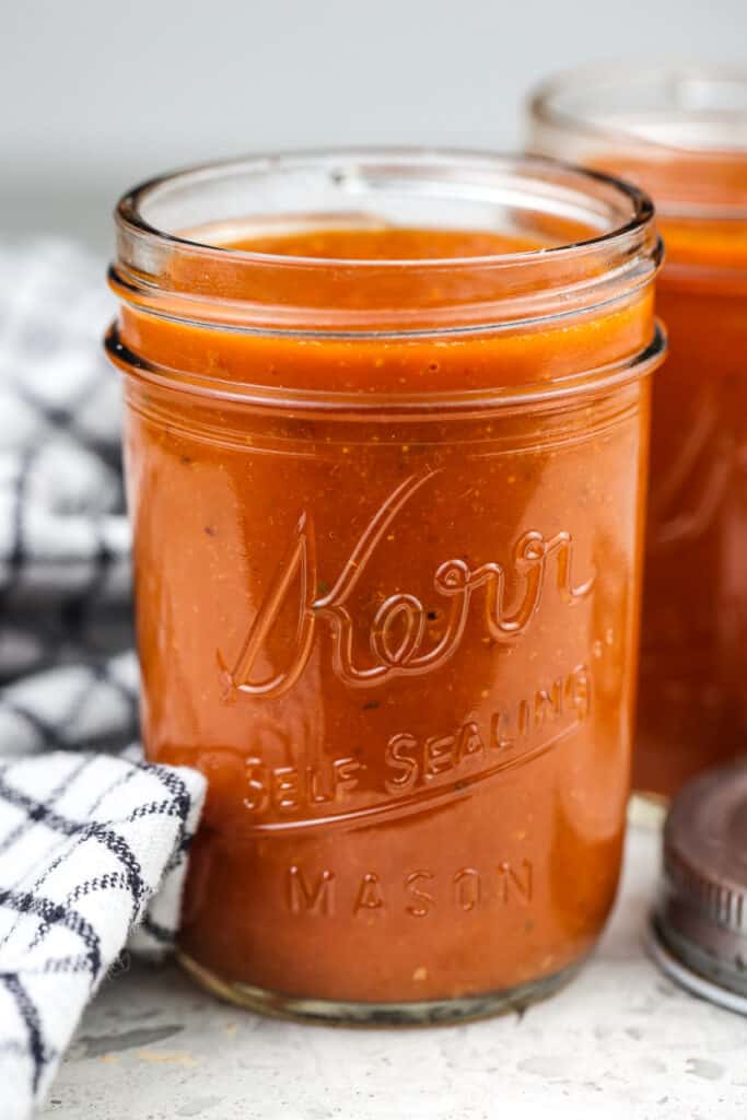 Picture of mason jar holding red enchilada sauce with second jar in the back next to two silver lids and a black towel.