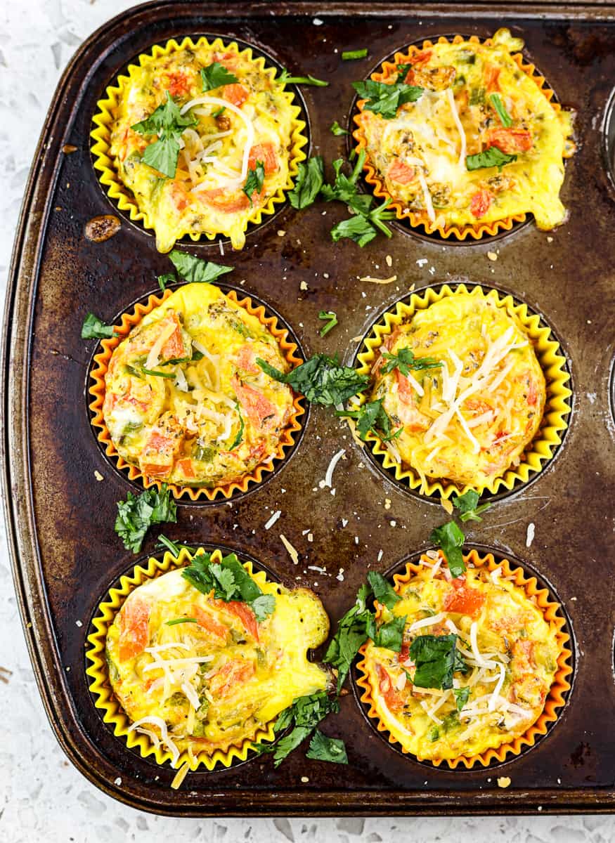 Old rusted muffin tin with 6 veggie and egg frittatas pictured on white marble background. 