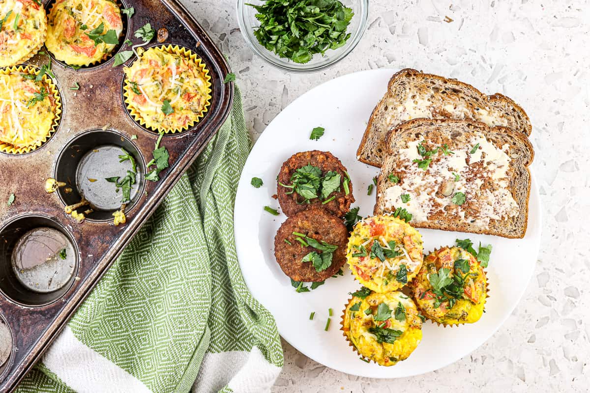 White marble backdrop with green towel and white plate filled with toast, sausage and eggs next to a muffin tin with a clear bowl of fresh herbs.