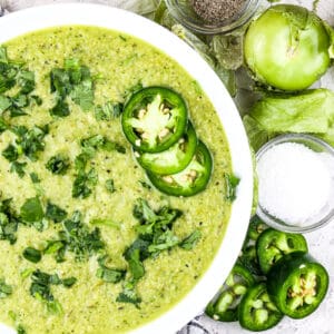 White bowl filled with hatch chile salsa verde topped with cilantro and jalapenos slices on a white backdrop.