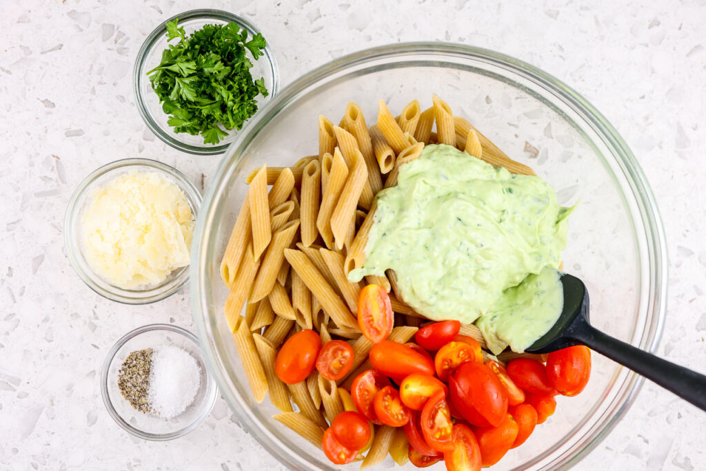 White tile backdrop with glass bowl filled with wheat pasta, red tomatoes, green sauce, and cheese, parsley and salt on the side.