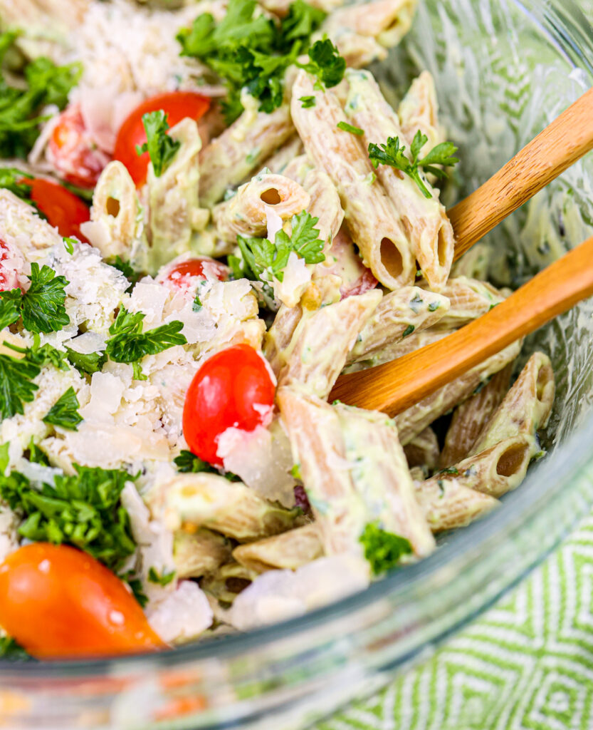 Glass bowl filled with brown penne pasta covered in green pesto sauce with red cherry tomatoes poking through.