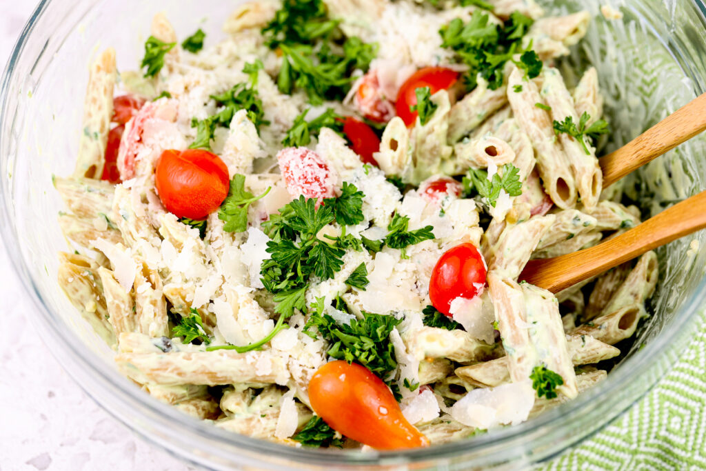 Glass bowl with wheat pasta covered in avocado pesto sauce and topped with chopped green parsley and halved red tomatoes.