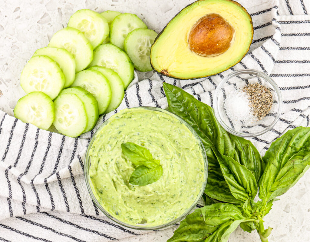 White tile backdrop with green pesto sauce in glass bowl under black towel with cucumbers, avocado and basil on the side.