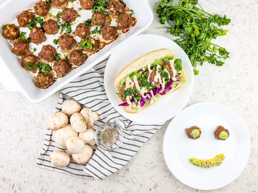 Baby meatballs in white casserole dish on the side with a hoagie roll stuffed with meatballs and greens in the center and a smiley face plate on the side with two meatballs for eyes and avocado mouth.