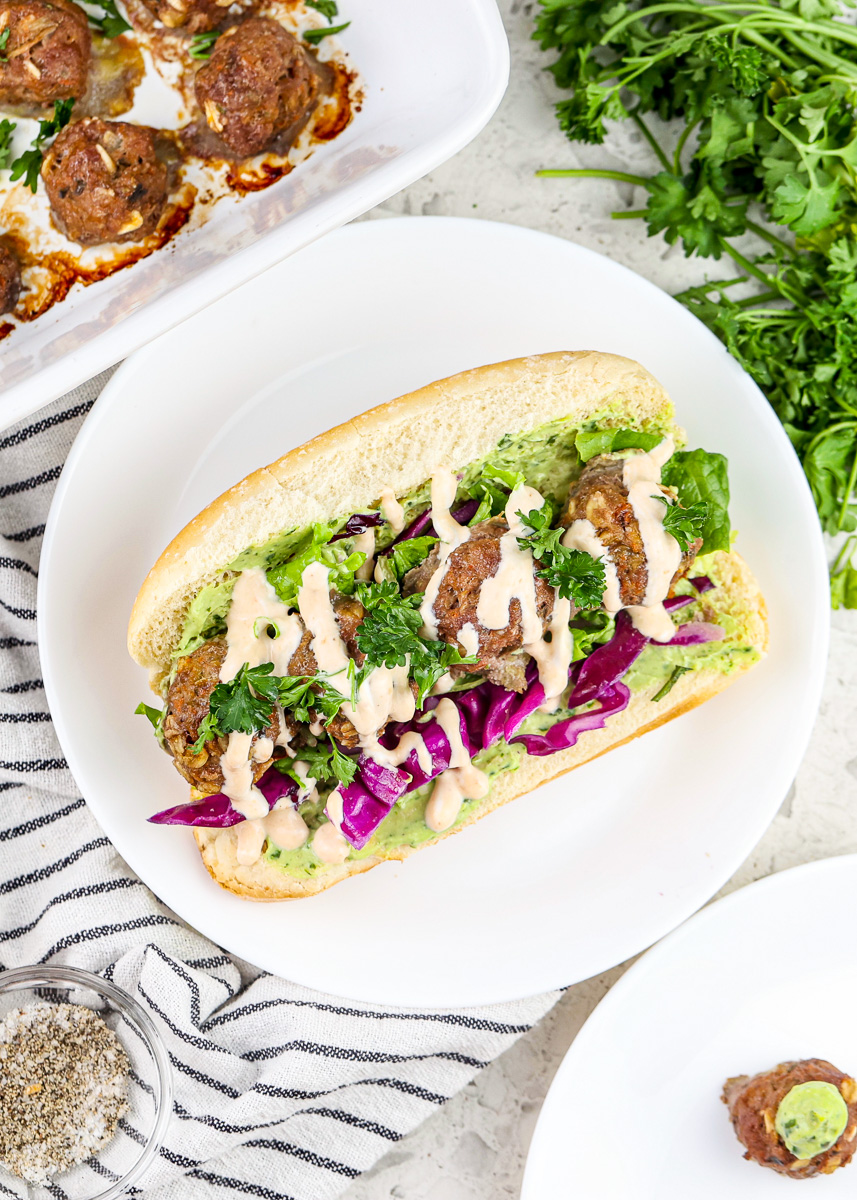 White plate with hoagie roll in center stuffed with baby meatballs, lettuce greens, and white sauce.