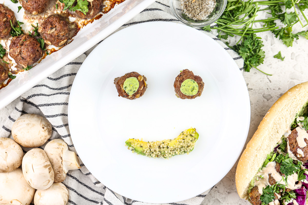 Picture of white plate with smiley face made out of two meatballs for eyes topped with green sauce and an avocado slice for a mouth.