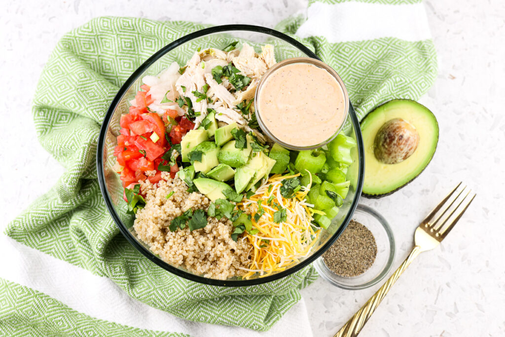 White tile with green napkin on top with salad bowl filled with greens, avocado, and tomato with dressing bowl on side.