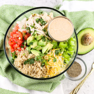 White tile with green napkin on top with salad bowl filled with greens, avocado, and tomato with dressing bowl on side.