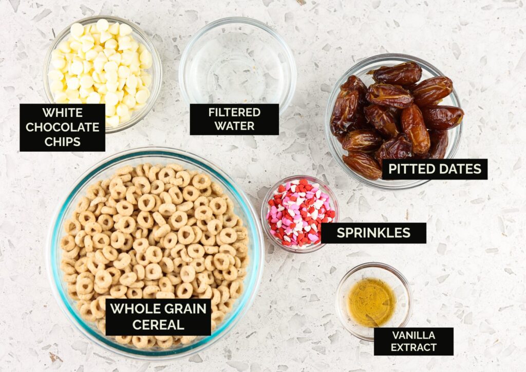 White marble backdrop with glass bowls of ingredients, including brown colored cheerios, chocolate chips, sprinkles, and dates to make Cereal Cake Pops.