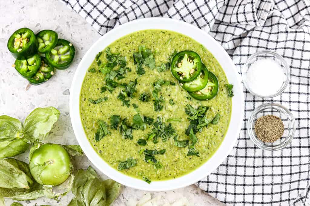 White bowl filled with hatch chile salsa verde topped with cilantro and jalapenos slices on a white backdrop.