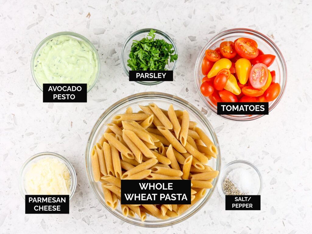 White tile backdrop with brown wheat pasta, cheese, tomatoes, and green pesto in glass bowls.