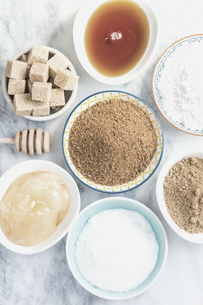 White background with bowls of brown sugar, cane. sugar, sugar cubes, and syrup.