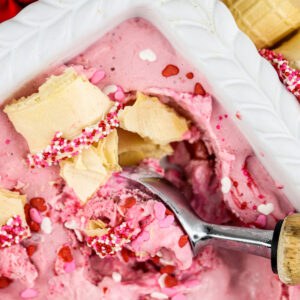 Picture of white pan with strawberry nice cream inside with crushed golden cake cones and a brown and silver ice cream scoop peaking out the side.