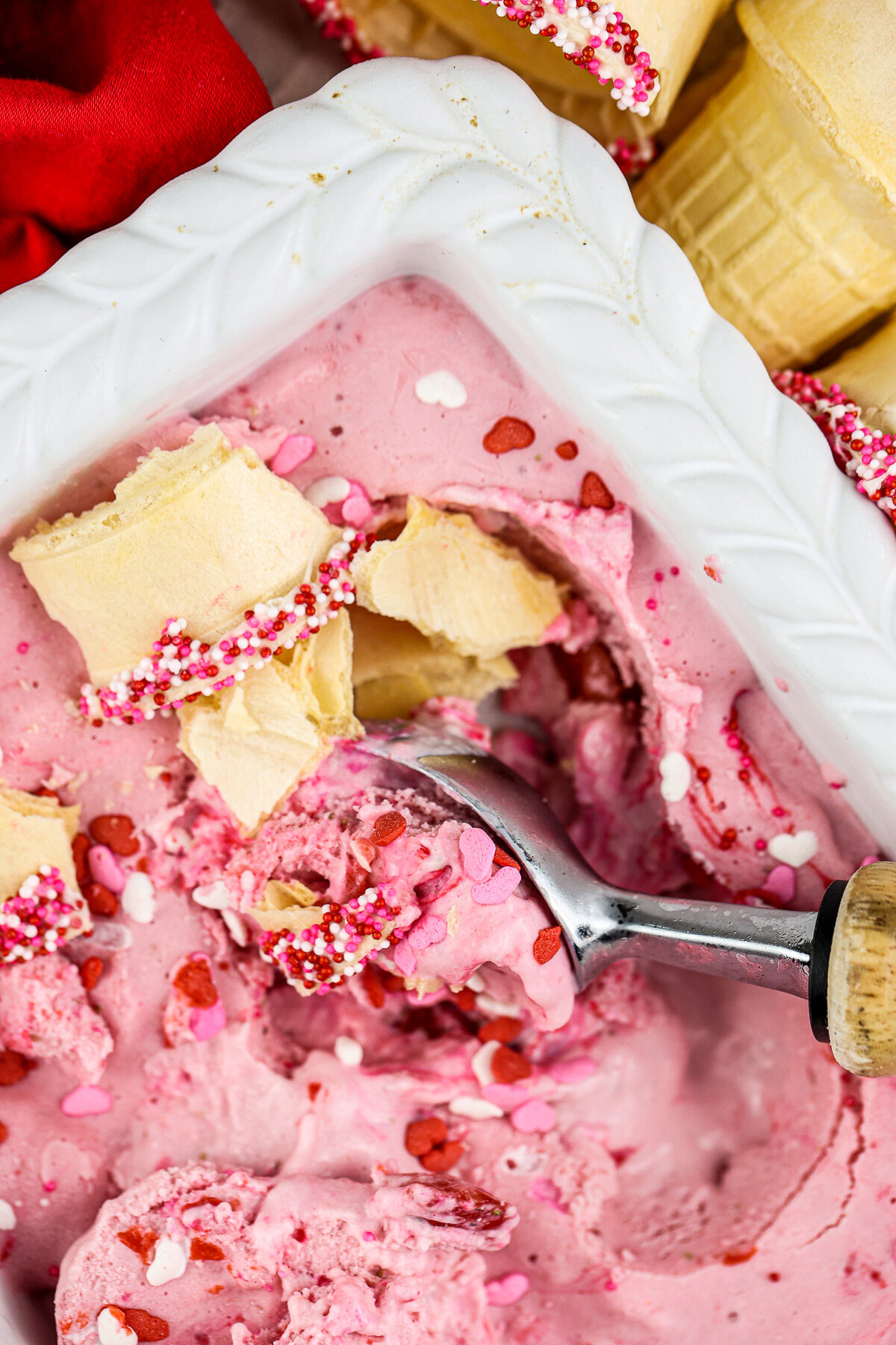 Picture of white pan with strawberry nice cream inside with crushed golden cake cones and a brown and silver ice cream scoop peaking out the side.