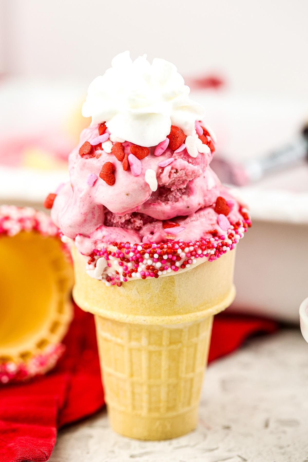 Picture of one cake cone filled with strawberry ice cream and topped with Valentines Day sprinkles and whipped cream.