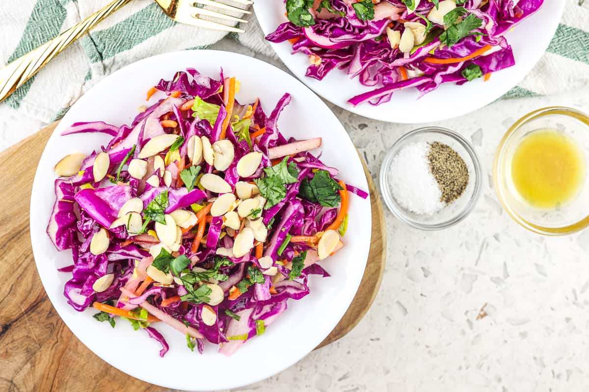 Two white plates with purple cabbage coleslaw on top with green napkin on a white marble backdrop.