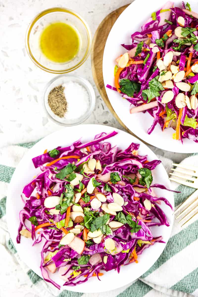 Two white plates with purple cabbage coleslaw on top with green napkin on a white marble backdrop.