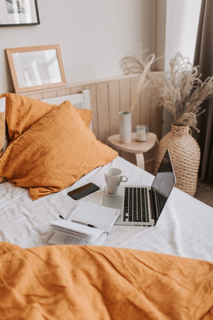 Silver and Black lap top on white bed with an empty white coffee mug on top of lap top. Open journal to the right of lap top. Orange pillows and comforter on the bed.