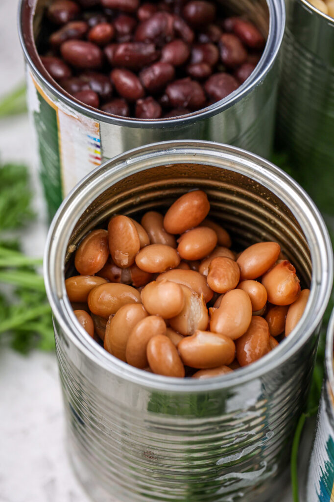 White tile backdrop with close up shot of pinto beans in open aluminum can and parsley in backdrop. 