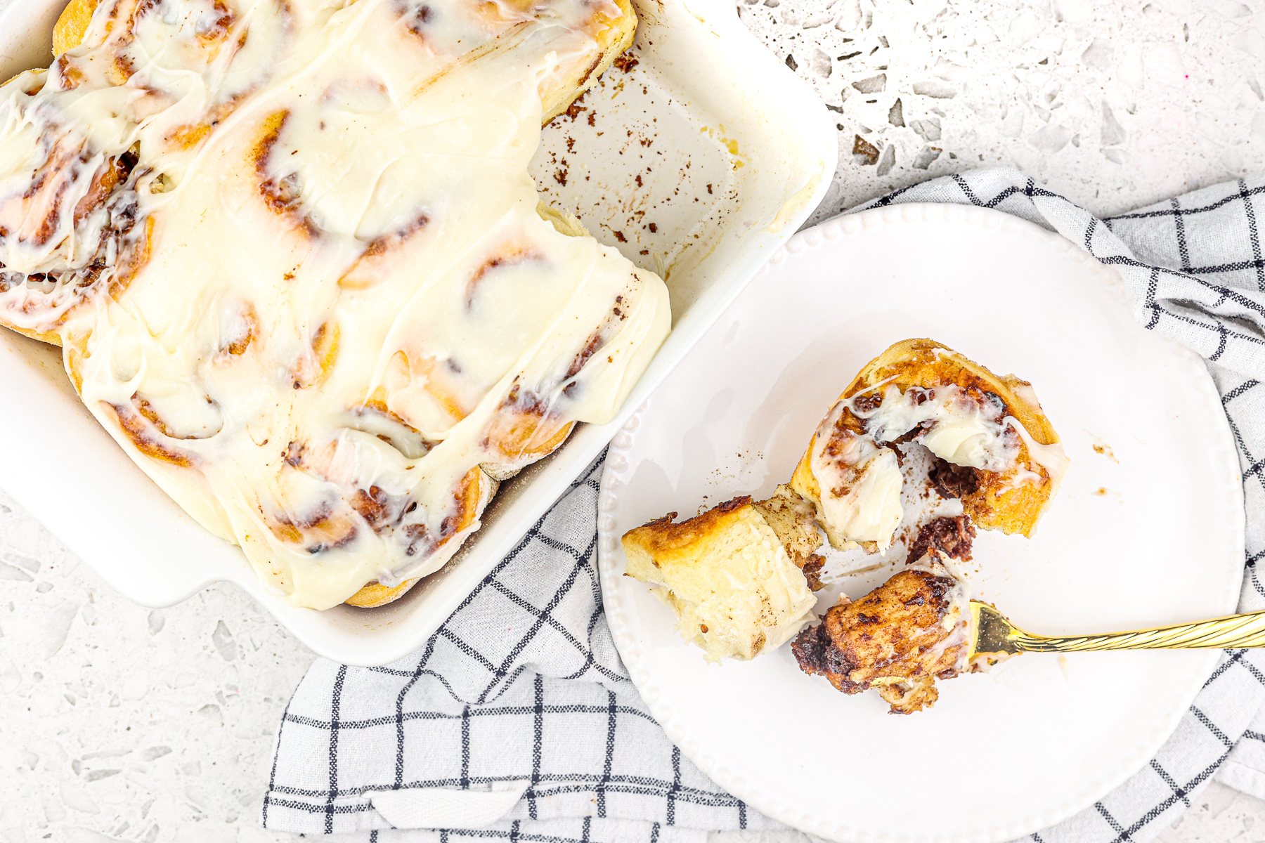 White tile backdrop with one cinnamon roll outside the white pan sliced open with a gold fork.