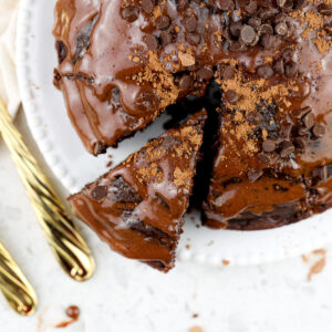 White tile background with chocolate cake slice coming out of a round cake with two gold forks.