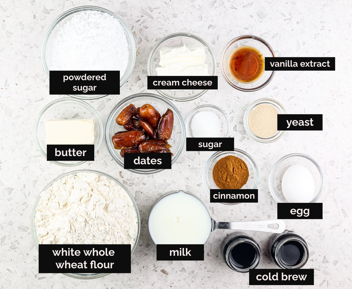 White tile backdrop with black text boxes on top of glass bowls holding ingredients. Flours, egg, dates, coffee, spices, and sugar are presented. 