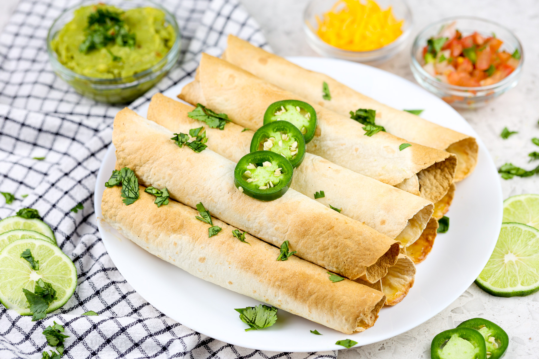 Black checkerboard napkin with white plate on top holding lightly browned taquitos with green garnish. 