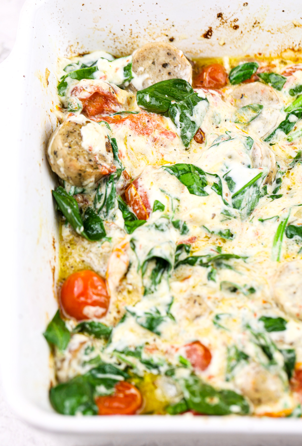 White backdrop with casserole dish filled with creamy white sauce, sausage, and tomatoes with cooked spinach.