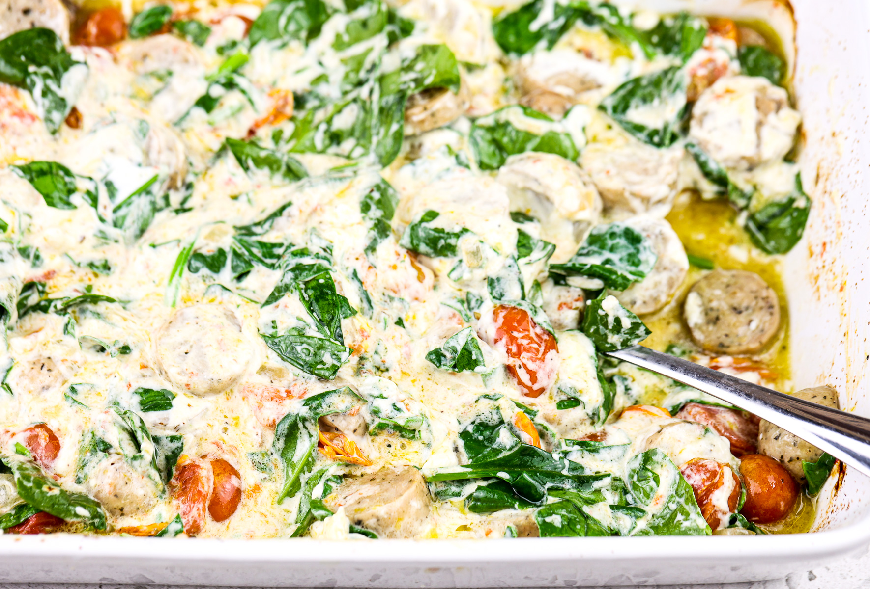 White backdrop with casserole dish filled with creamy white sauce, sausage, and tomatoes with cooked spinach.