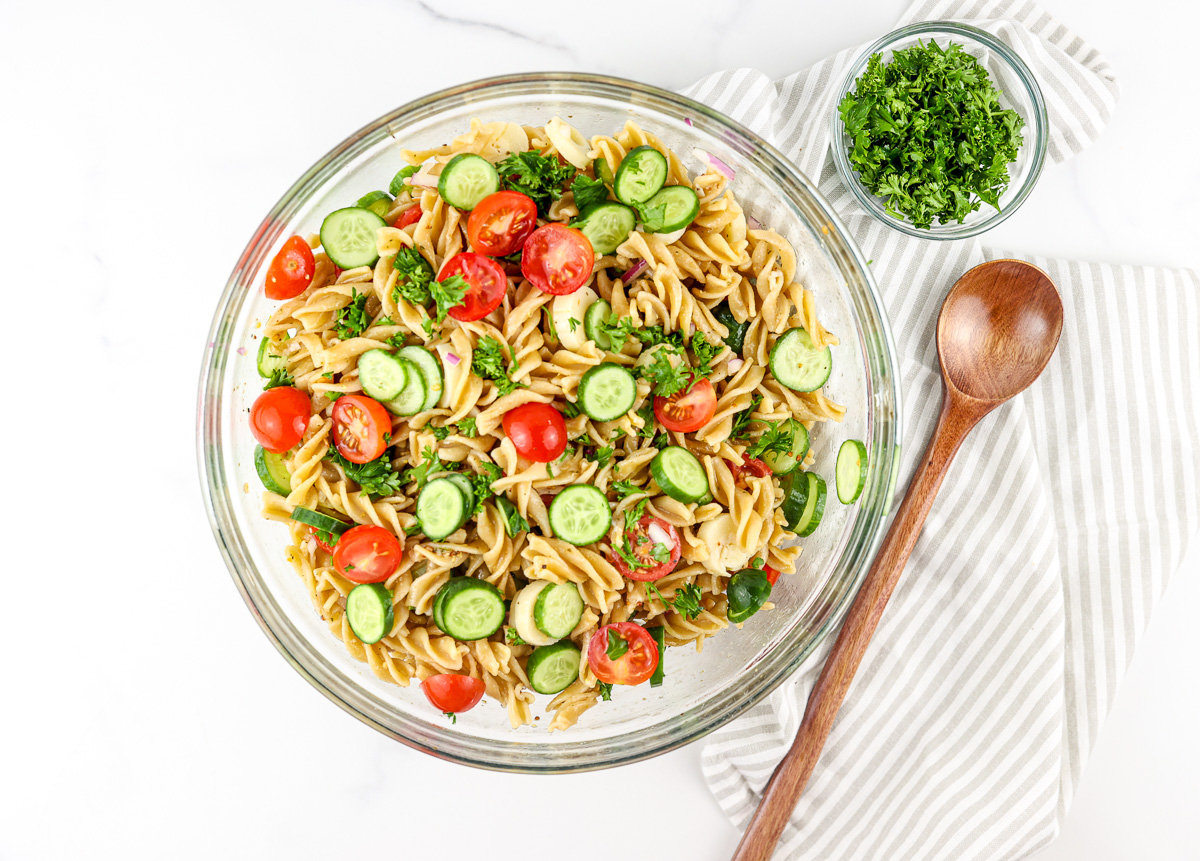 Pasta salad in white bowl with wooden spoon.