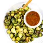 White plate and backdrop with green roasted brussel sprouts and red miso dressing in bowl.