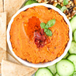 Image of red dip in bowl with cucumbers and pita around.