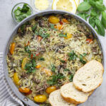 Styled shot of orzotto with grey towel holding saute pan and bread sliced on the pasta.