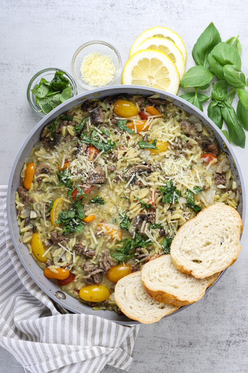 Styled shot of orzotto with grey towel holding saute pan and bread sliced on the pasta.